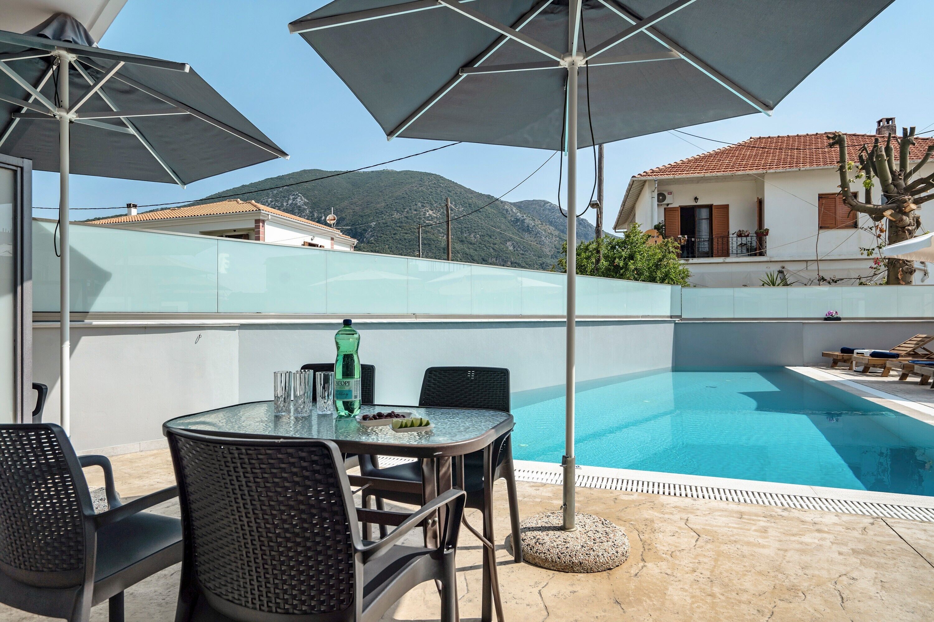 Un espace aménagé au bord de la piscine, avec une table en verre, des chaises, des parasols et une vue sur une montagne.