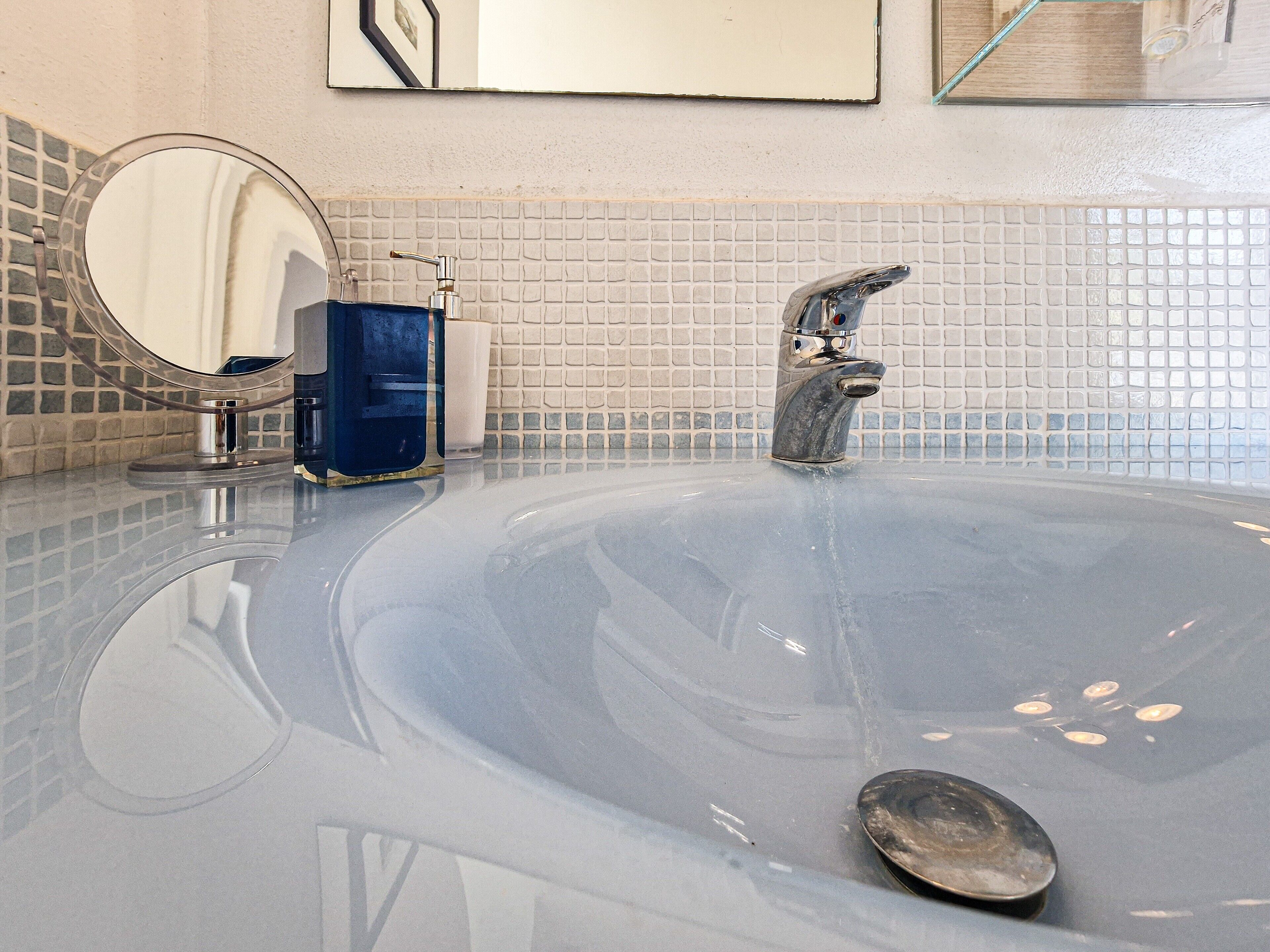 A bathroom with a large, clear bathtub, a round mirror, and a soap dispenser.