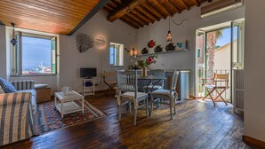 A room with wooden flooring, a dining table with chairs, a sofa, a TV, and a window with a view.