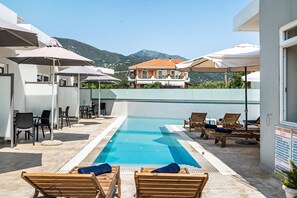 A pool area with lounge chairs and umbrellas, a clear view of mountains, and a building with a balcony in the background.