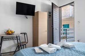 Une chambre d’hôtel avec un lit, un téléviseur fixé au mur, une armoire en bois, une petite table avec des fruits, une chaise et une vue sur l’extérieur par une porte ouverte.