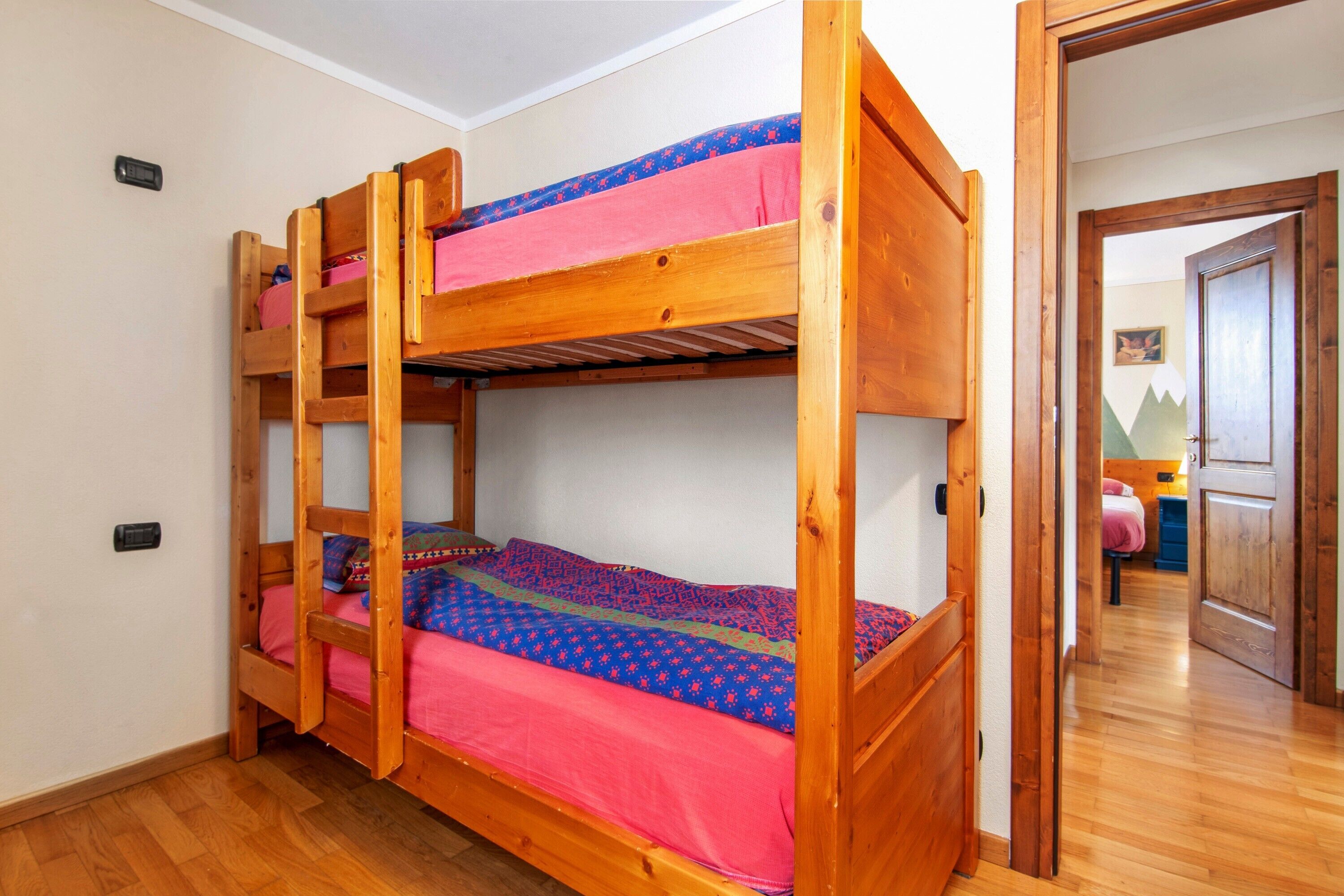 A bunk bed with a wooden frame, a lower bunk with a pink mattress, and a top bunk with a blue mattress. There is a door leading to another room.