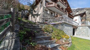 Un immeuble à plusieurs étages, avec des marches en pierre menant à un balcon en bois, entouré de verdure et dominé par un paysage montagneux.