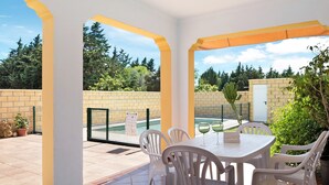 Um alpendre coberto com piscina, cadeiras brancas e uma mesa com copos.