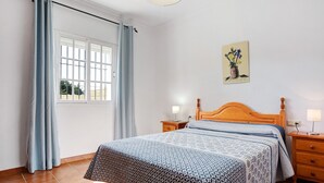Quarto com cama, mesa de cabeceira, cabeceira de madeira, janela com cortinas e um quadro na parede.