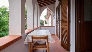 Ein überdachter Patio-Bereich mit Tisch und Stühlen, Holzläden und Blick auf ein Gebäude im Hintergrund.