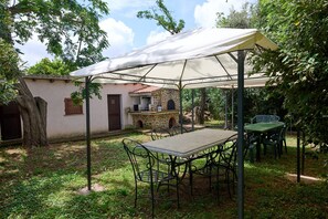 Eine überdachte Terrasse mit Tisch und Stühlen, ein Steinhaus mit Schornstein und ein Baum.