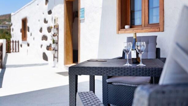 A wicker table with two wine glasses and a bottle, set against a white building with a wooden window frame. - Rural Holiday Home 'Alto Prado' with Sea View, Private Terrace and Wi-Fi (Valverde)