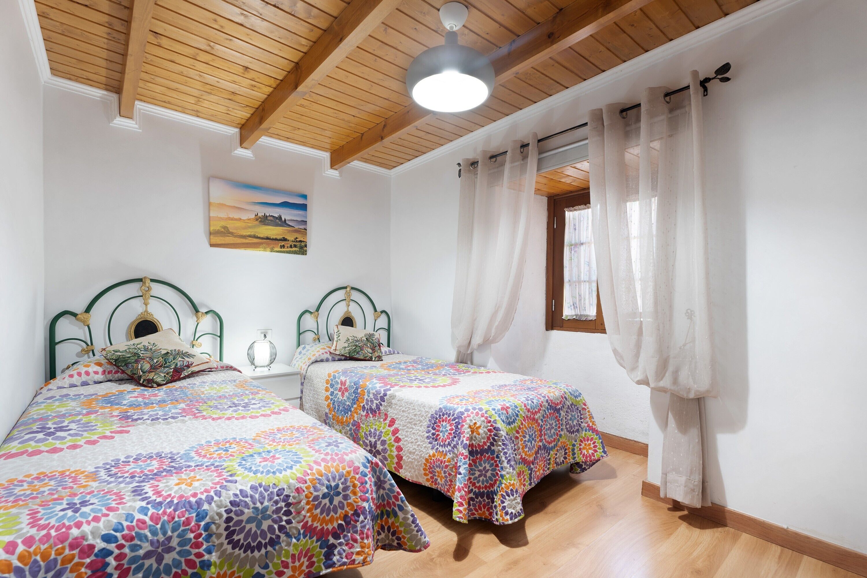 A bedroom with two beds, wooden ceiling beams, a ceiling fan, a window with curtains, and a painting on the wall.