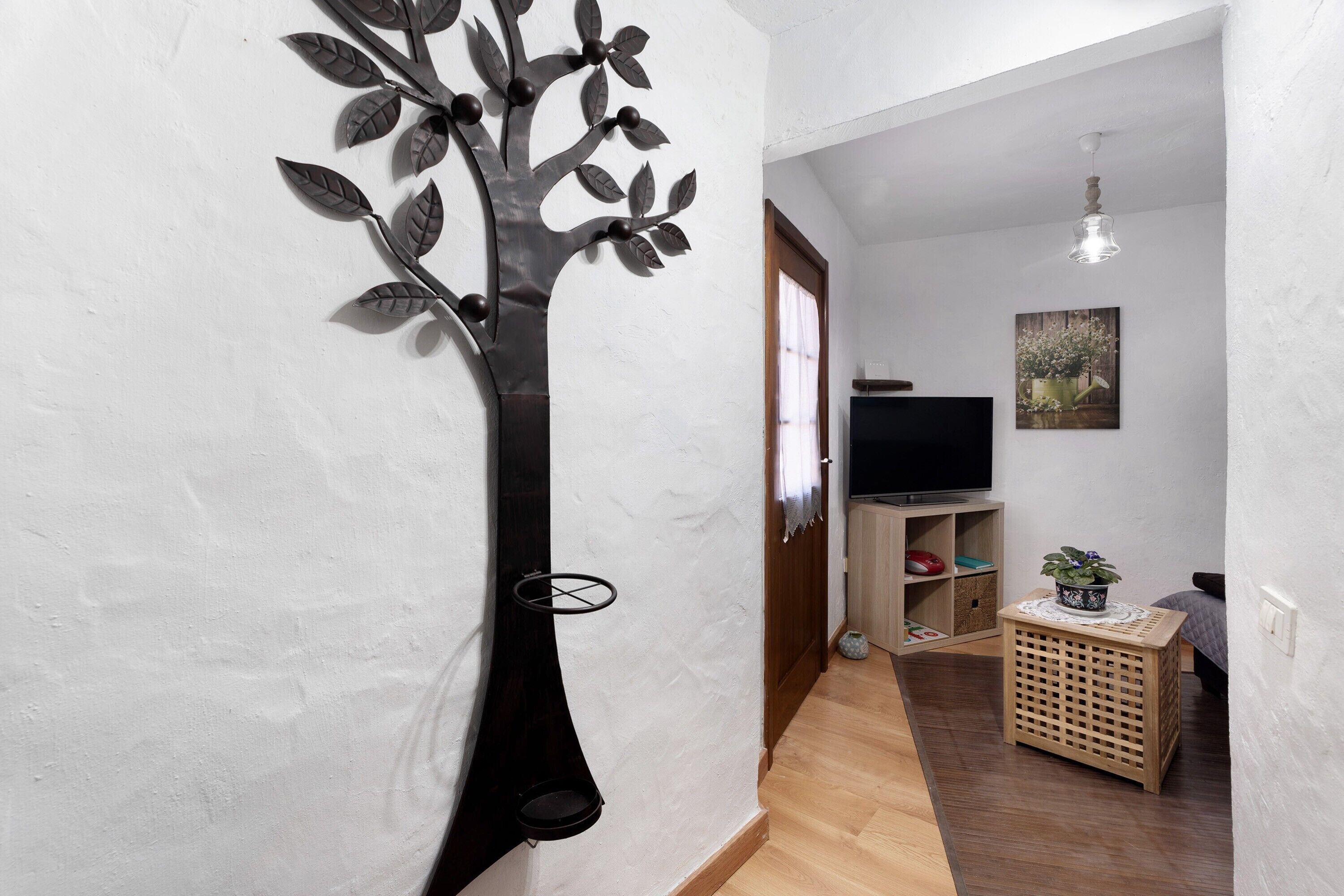 A living room with a TV, a decorative tree wall piece, a wooden floor, a small wooden side table with a plant, and a painting on the wall.