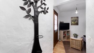 A living room with a TV, a decorative tree wall piece, a wooden floor, a small wooden side table with a plant, and a painting on the wall.
