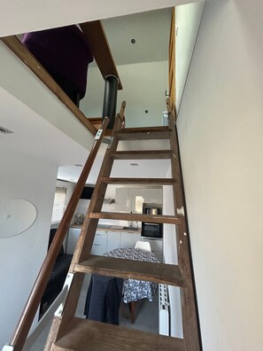 Un escalier intérieur en bois, doté d’une rampe, mène à une pièce où l’on aperçoit un coin cuisine à l’arrière-plan.