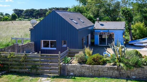 A blue metal building with a sloped roof, a small attached structure with a tiled roof, and a stone wall with plants. - Petit duplex proche mer (Plouhinec)