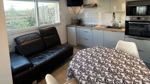 A compact kitchen with a microwave, oven, and a small refrigerator. There is a sofa and a dining table with a patterned tablecloth. The room has a large window and a flat-screen TV mounted on the wall. - Petit duplex proche mer (Plouhinec)