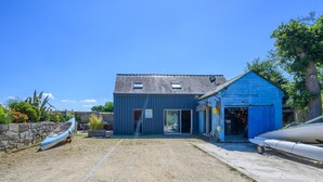 A building with a blue exterior, a covered outdoor area, and a boat docked outside. - Petit duplex proche mer (Plouhinec)