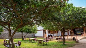 Un aula al aire libre con mesas y sillas, rodeada de árboles y un edificio con ventanas arqueadas.