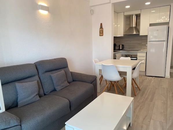 A modern living room with a grey sofa, a white table, and a kitchen area with white cabinets and a white refrigerator.