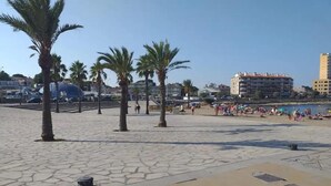 Ein gepflasterter Promenadenspaziergang mit Palmen, ein Strand voller Menschen und Gebäude im Hintergrund.