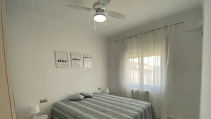 Ein Schlafzimmer mit einem Bett, einem Deckenventilator, einem Nachttisch, einem Fenster mit Vorhängen und Wandbildern.