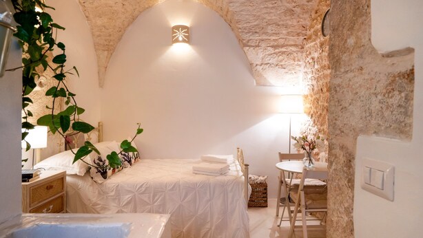 Ein gemütliches Schlafzimmer mit Steinwand, einem Bett mit weißer Bettdecke, einem kleinen Tisch mit einer Vase und einem Sessel.