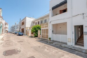 Une rue résidentielle bordée d’immeubles blancs, avec une voiture bleue garée sur le côté et un arbre au premier plan.