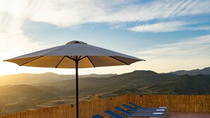 Una terraza de madera con una sombrilla grande, sillas de lounge azules y un paisaje montañoso al fondo.