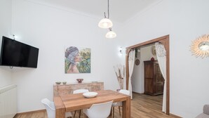 A dining area with a wooden table, white chairs, a TV mounted on the wall, and a painting above the table. - Holiday Home 'Maison Chloe' close to the Beach with Terrace, Garden & Wi-Fi (La Maddalena)