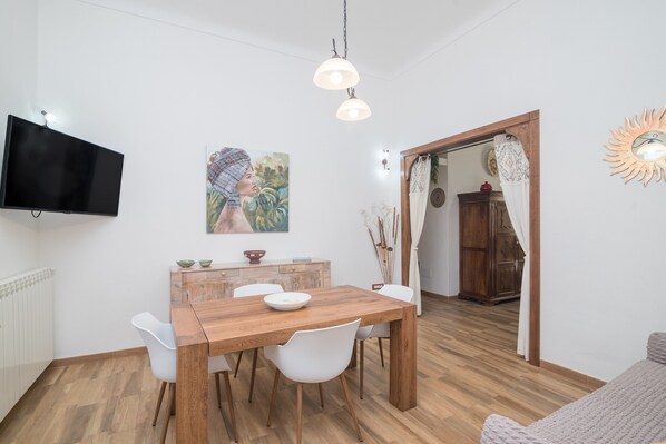 Un comedor con mesa de madera, sillas blancas, televisor montado en la pared y un cuadro sobre la mesa.