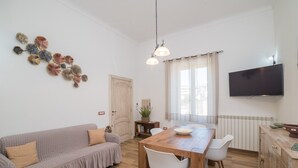 A living room with a sofa, dining table, chairs, a TV, and a decorative wall piece. - Holiday Home 'Maison Chloe' close to the Beach with Terrace, Garden & Wi-Fi (La Maddalena)