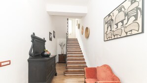 A hallway with a staircase, a dark wooden dresser, a red upholstered chair, and wall-mounted artwork.