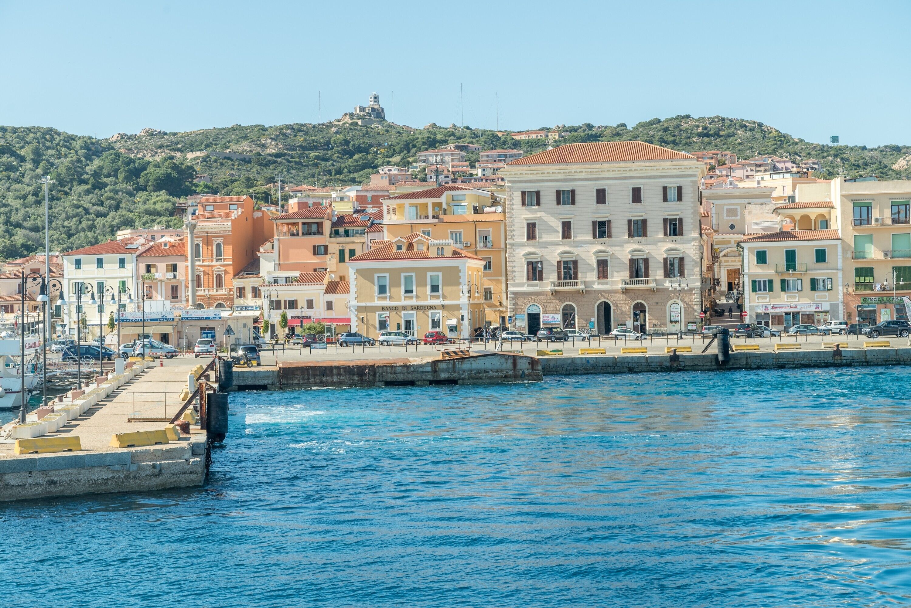 A coastal town with buildings along the waterfront, a prominent hill in the background, and a clear blue sky.