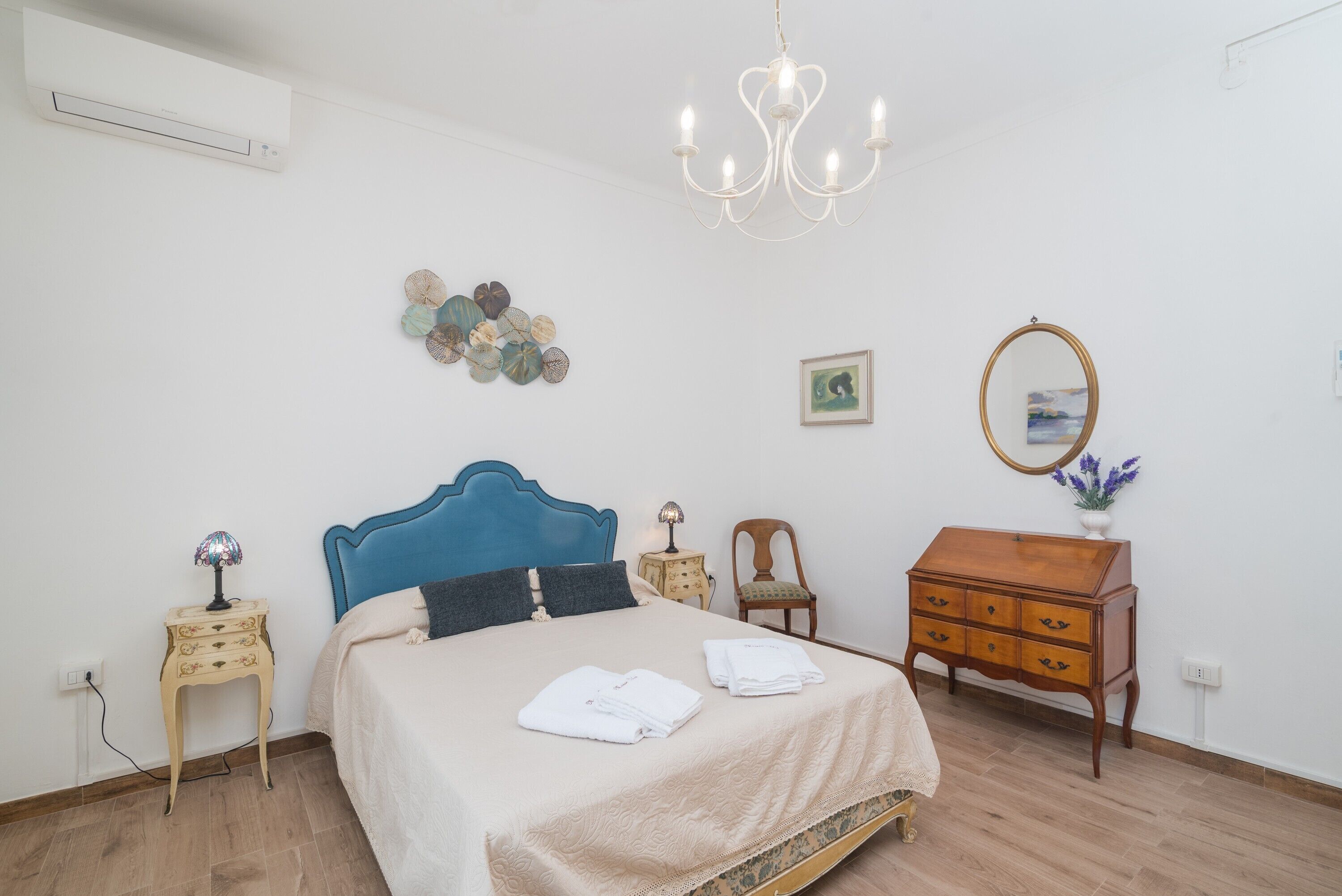 Een slaapkamer met een bed, nachtkastjes, een kaptafel, een kroonluchter, wanddecoratie en een airconditioning.