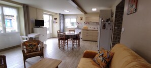 Una sala de estar con un sofá y una mesa de comedor con sillas, una cocina con un refrigerador y una ventana, y un televisor sobre un mueble.