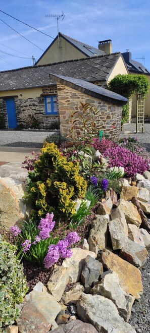 Una casa de piedra con puerta azul, chimenea y jardín con plantas y rocas diversas.