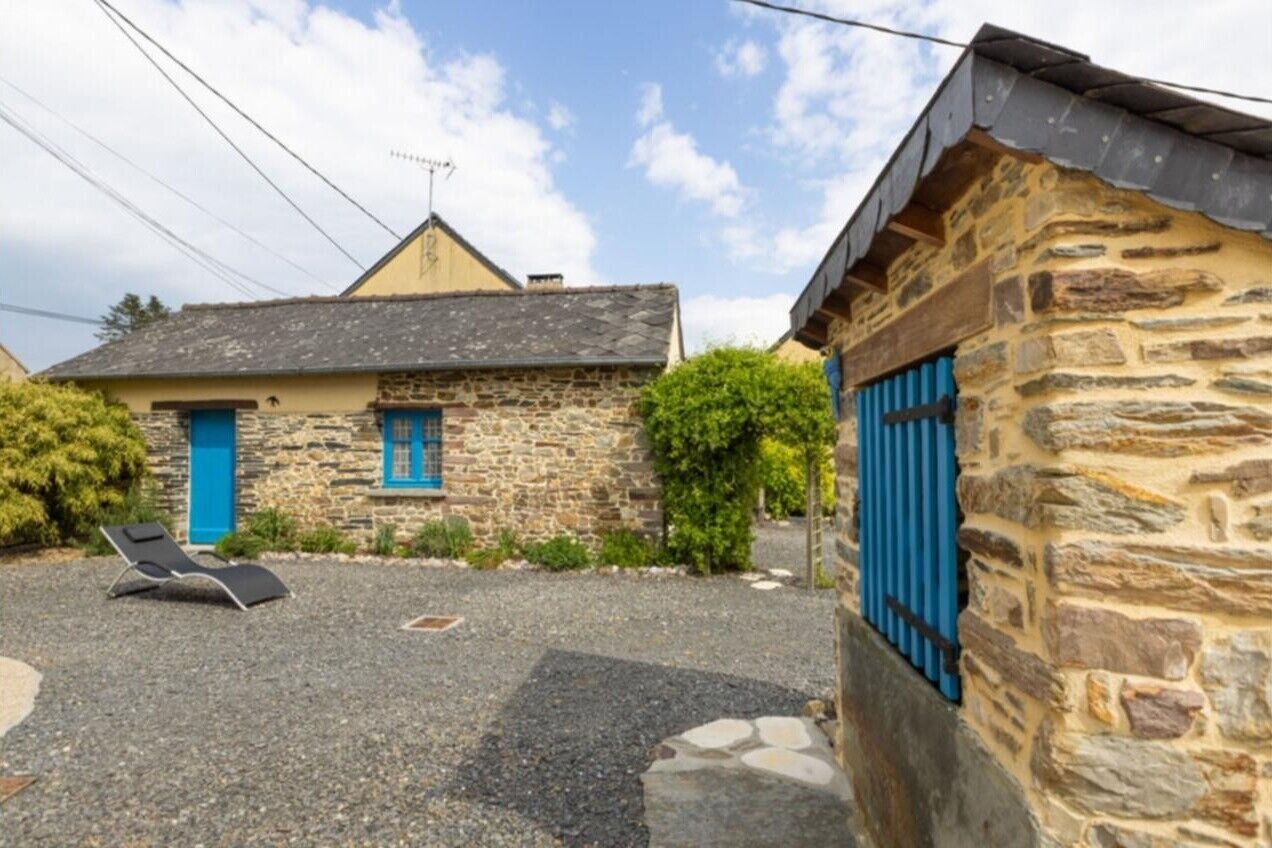 A stone building with a blue door and shutters, a gravel courtyard, and a lounge chair.