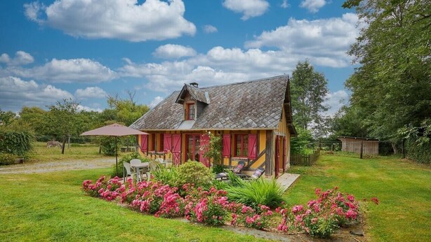 A rustic house with a sloped roof, a porch with a table and chairs, and a garden with pink flowers.