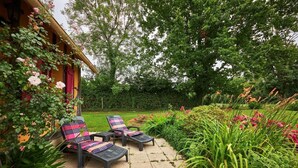 A patio with two lounge chairs, a garden with various plants and flowers, and a wooden building with a red door.