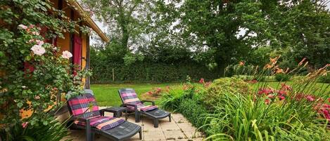 A patio with two lounge chairs, a garden with various plants and flowers, and a wooden building with a red door.