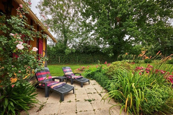Un patio con due poltrone, un giardino con diverse piante e fiori, e un edificio in legno con una porta rossa.