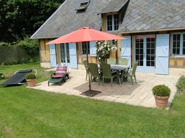 Une maison avec un patio, une table entourée de chaises, un parasol rouge et un petit jardin.