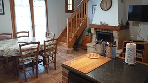 Un comedor con mesa y sillas de madera, una encimera de cocina con tabla de cortar, una chimenea y un reloj en la pared.