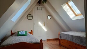 Une petite chambre mansardée avec deux lits simples, une porte blanche et des fenêtres de toit.