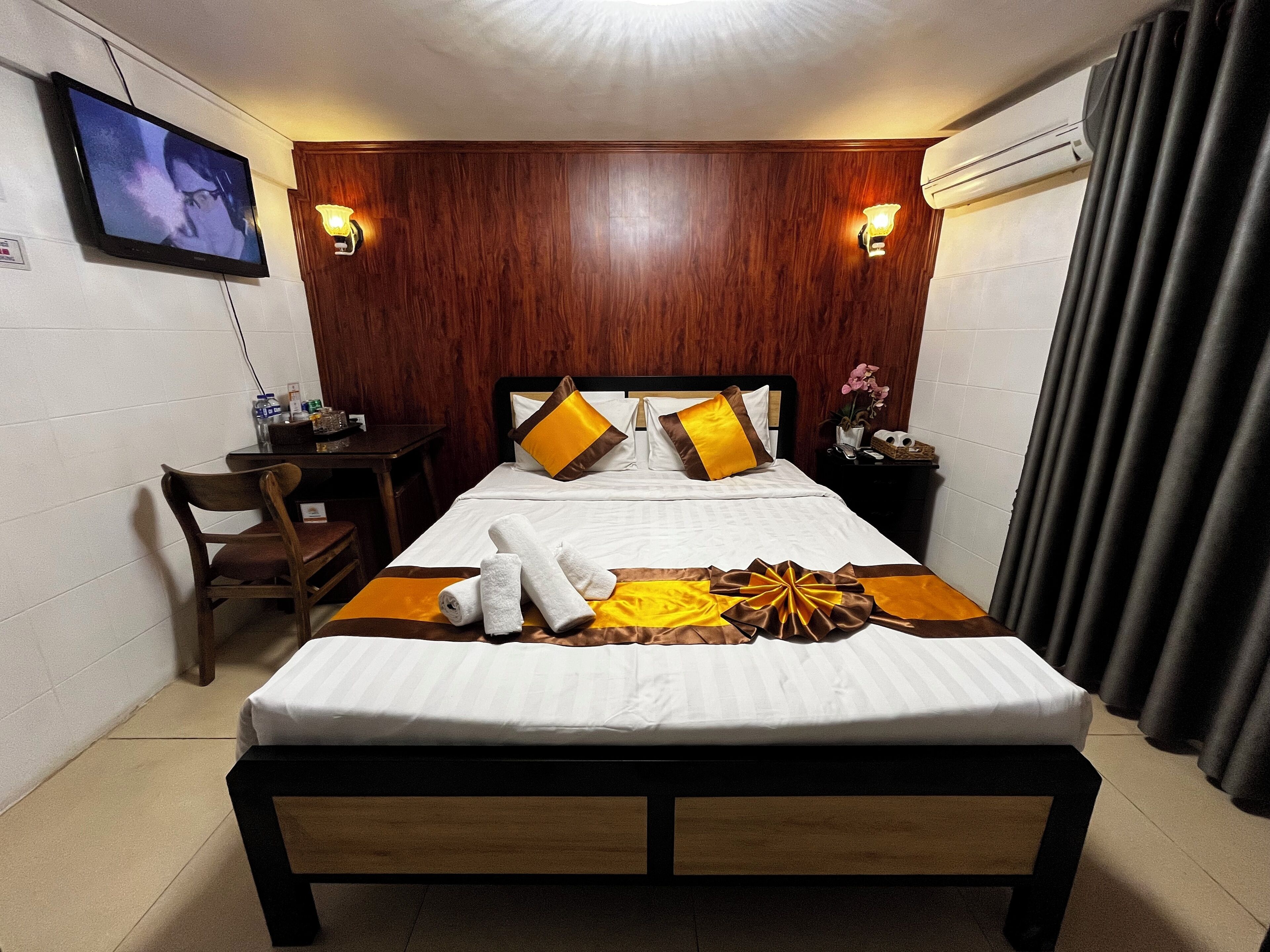 A hotel room with a wooden headboard, a bed with white bedding and decorative pillows, a bedside table with a lamp, a chair, and a TV mounted on the wall.