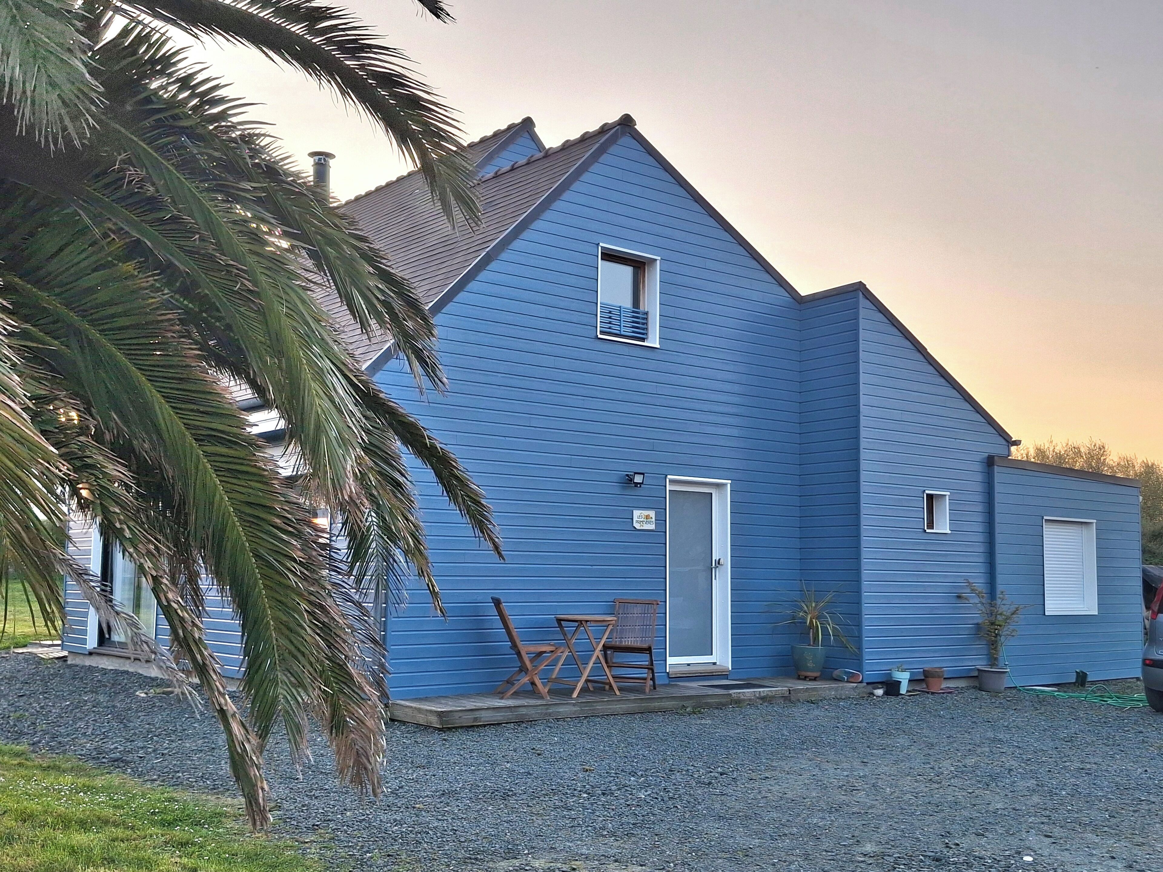 A blue house with a sloped roof, a small porch with a chair and a table, and a gravel driveway.