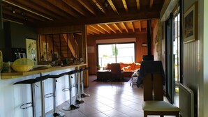 A kitchen with a bar area, a dining area with a sofa, and a large window. - Maison en Bois sur Grand Jardin et à 5 Minutes de la Plage (Plobannalec-Lesconil)