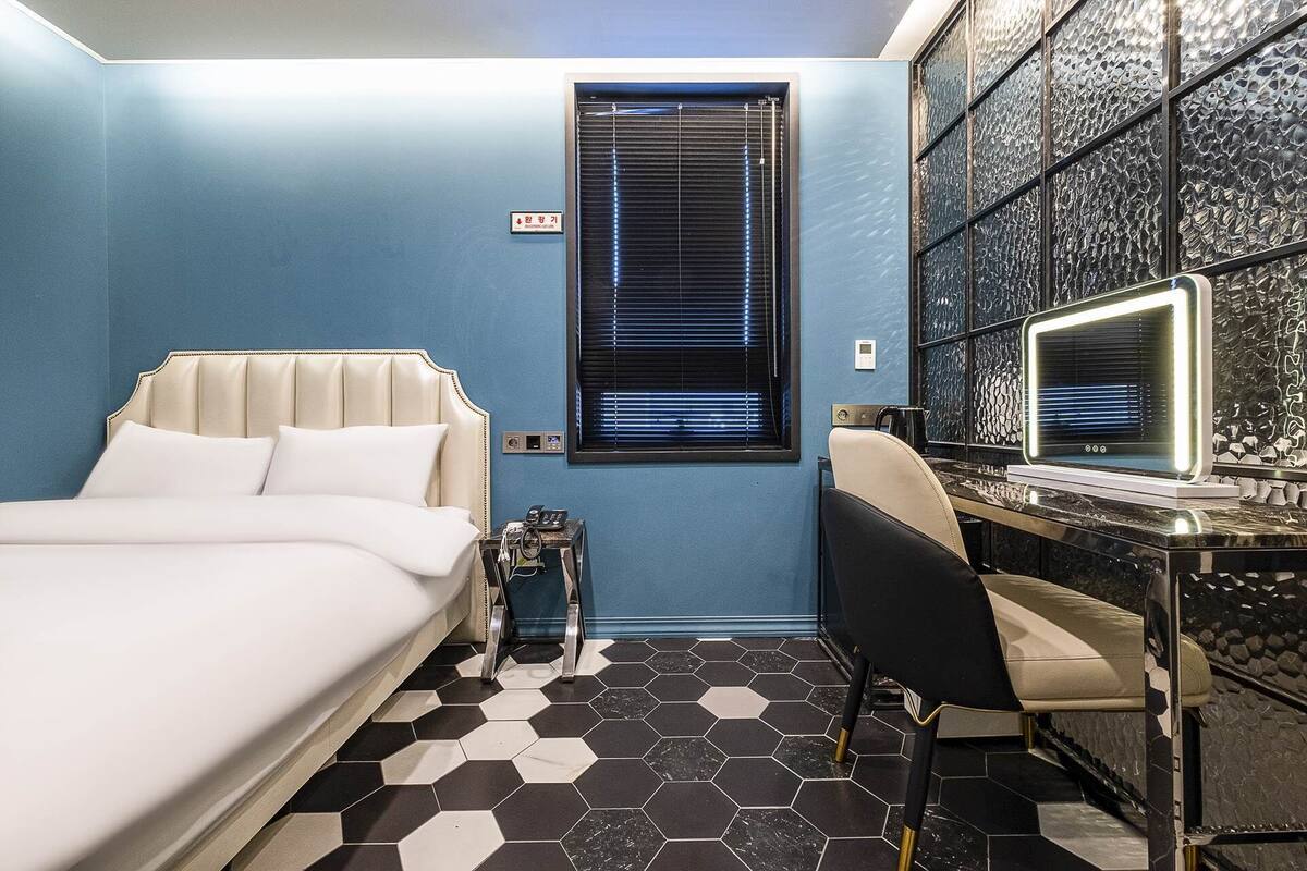 Habitación de hotel con cama, escritorio con computadora, silla, mesita pequeña y ventana con persianas.