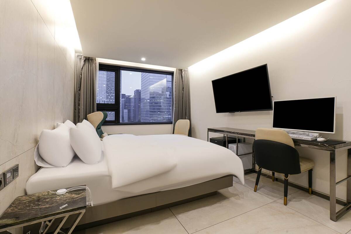 Una habitación de hotel moderna con una cama grande, un escritorio con computadora, un televisor y vistas a la ciudad a través de la ventana.