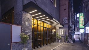 Entrada de un hotel con fachada moderna, amplias puertas de vidrio y un cartel con el nombre y la dirección.