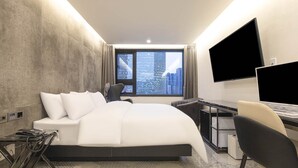 Una habitación de hotel moderna con una cama grande, un escritorio, una silla, un televisor de pantalla plana y vista a la ciudad desde la ventana.