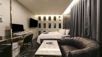 A modern hotel room with a bed, a desk with a computer, a TV, and a leather armchair.